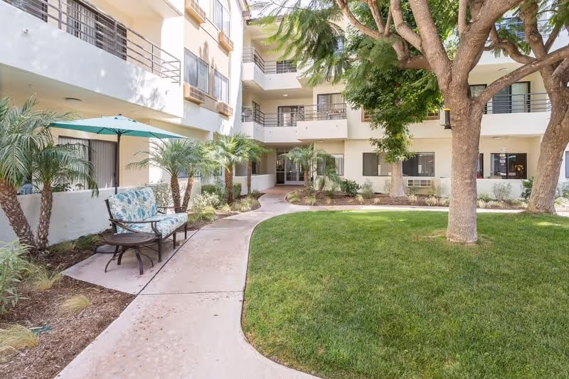 A landscaped courtyard with a curved paved path, grassy lawn, trees, patio seating under an umbrella, and surrounding apartment balconies.