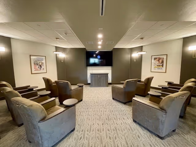 A small theater room with two rows of comfortable recliner chairs facing a wall-mounted flat screen TV. The room has patterned carpet, soft lighting fixtures on the walls, and framed movie posters on either side of the TV.