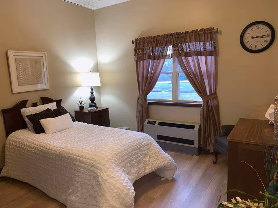 A neatly furnished single-occupancy bedroom with a bed, nightstand and lamp, window with curtains, dresser and wall clock.