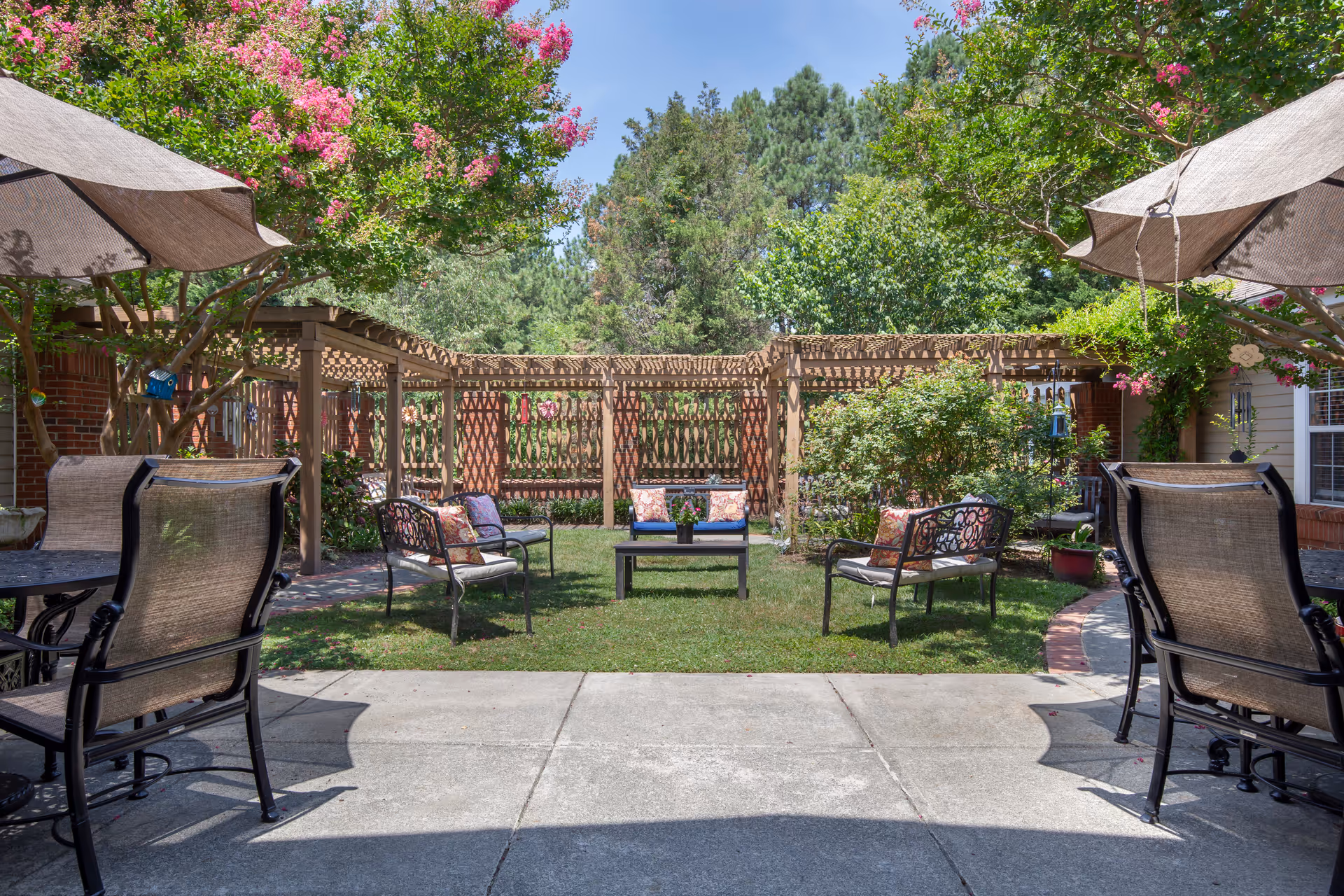 Sunny outdoor courtyard with patio tables and chairs, umbrellas, a central seating area, and a wooden pergola surrounded by greenery.