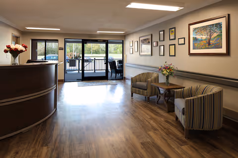 Reception lobby with a curved front desk, two upholstered chairs and a small table, framed artwork on the wall, and glass entrance doors leading outside.