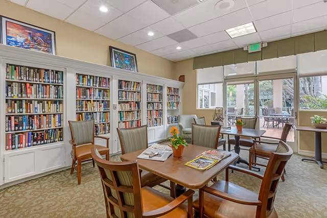 A cozy reading and sitting area in a senior living facility with bookshelves filled with books along the wall, several wooden chairs with cushions around tables, potted plants on the tables, and large windows and glass doors leading to an outdoor patio.