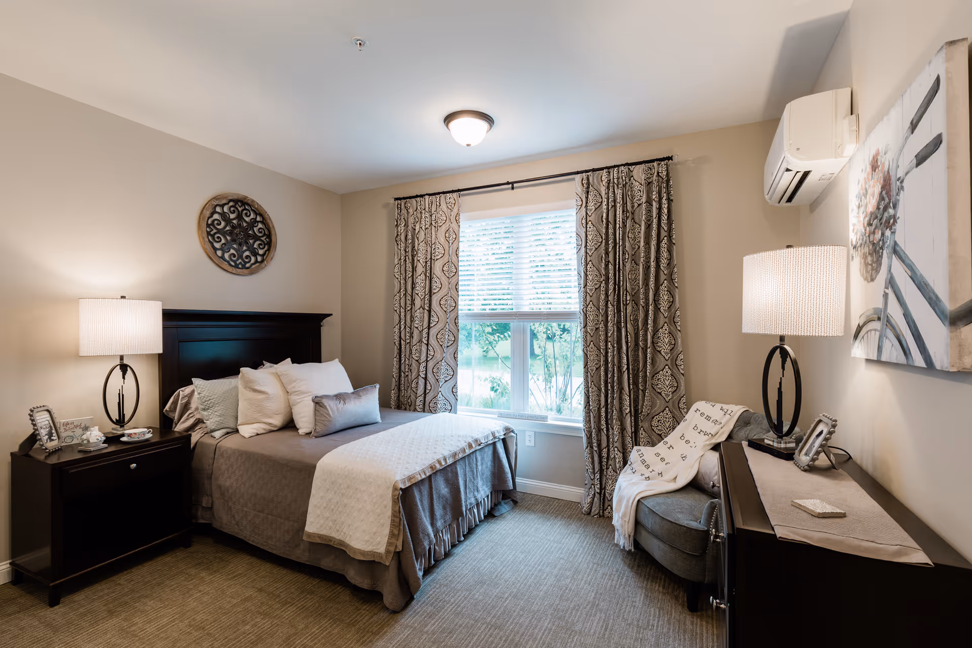 A well-lit bedroom with a made bed, nightstands and lamps, patterned curtains over a window, and a small armchair.