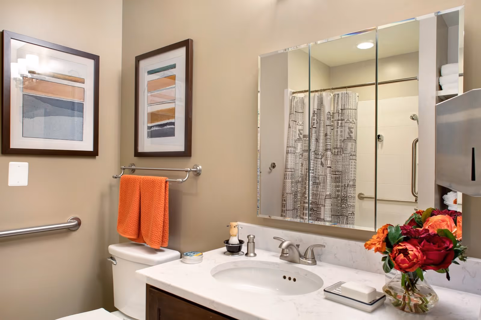 A clean and well-lit bathroom featuring a white toilet with an orange towel hanging on a silver towel rack above it. Above the toilet are two framed abstract art pieces. A white countertop with a built-in sink and silver faucet is visible, with a soap dish and a small decorative item on it. A large mirror with three panels is mounted above the sink, reflecting a shower area with a curtain that has a cityscape design. A vase with red and orange flowers is placed on the countertop. The walls are painted beige.