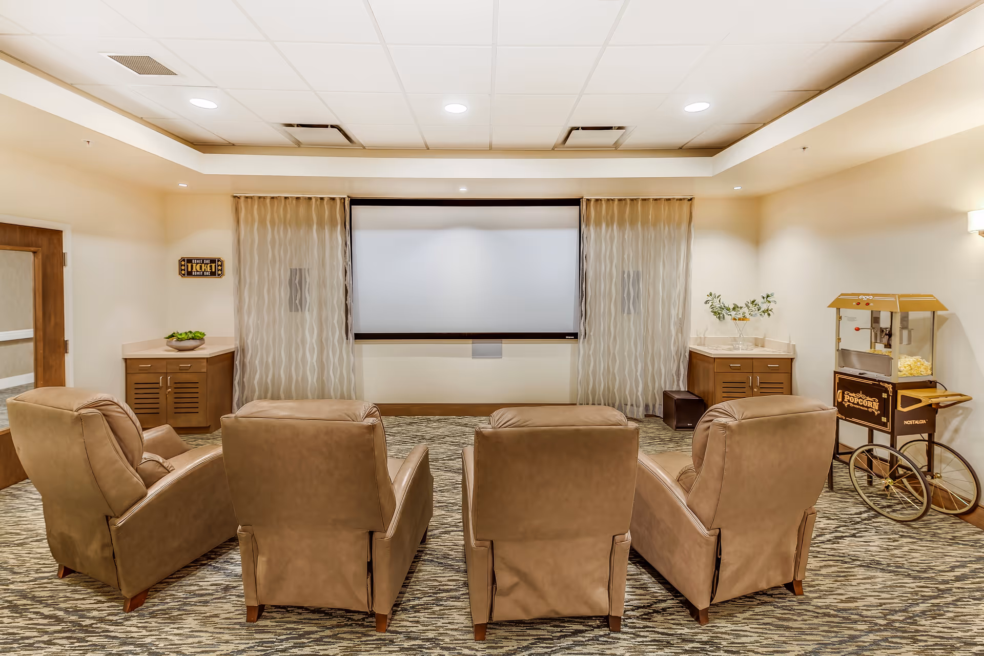A cozy media room with four brown recliner chairs facing a large blank projector screen. The room has beige walls, patterned carpet, and soft lighting. To the right, there is a vintage-style popcorn machine on a cart. On the left side, there is a wooden door and a small cabinet with a decorative bowl on top. Sheer curtains frame the projector screen.