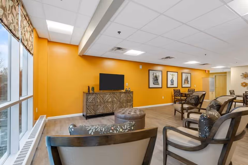 A bright and spacious senior living common area with large windows on the left letting in natural light. The walls are painted a warm yellow-orange color. There is a wooden cabinet with a flat-screen TV mounted above it against the wall. Several comfortable chairs with patterned cushions are arranged around a round ottoman in the center. In the background, there are tables and chairs for dining or socializing, and framed artwork hangs on the wall.