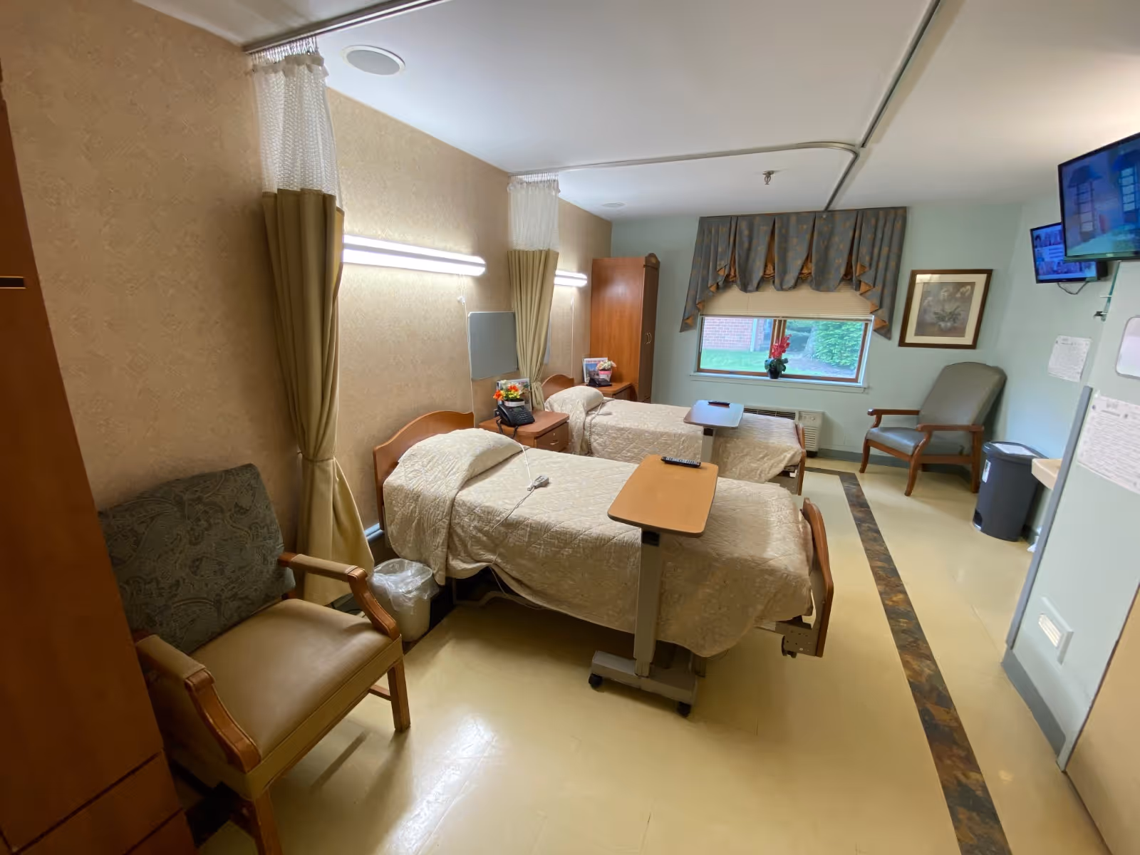 Two hospital-style beds with overbed tables, chairs, and a window in a shared senior care room.