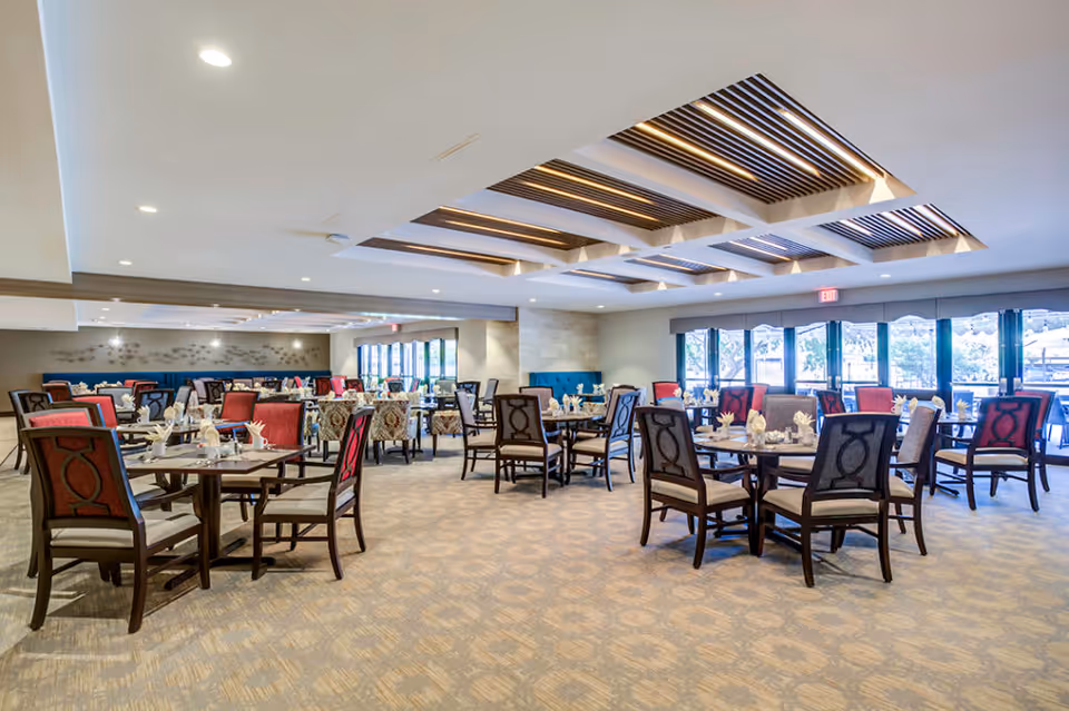 Spacious dining room with multiple round tables and chairs arranged neatly. The chairs have a mix of red and patterned upholstery. The room features large windows allowing natural light to enter, and the ceiling has modern recessed lighting with wooden slats design.