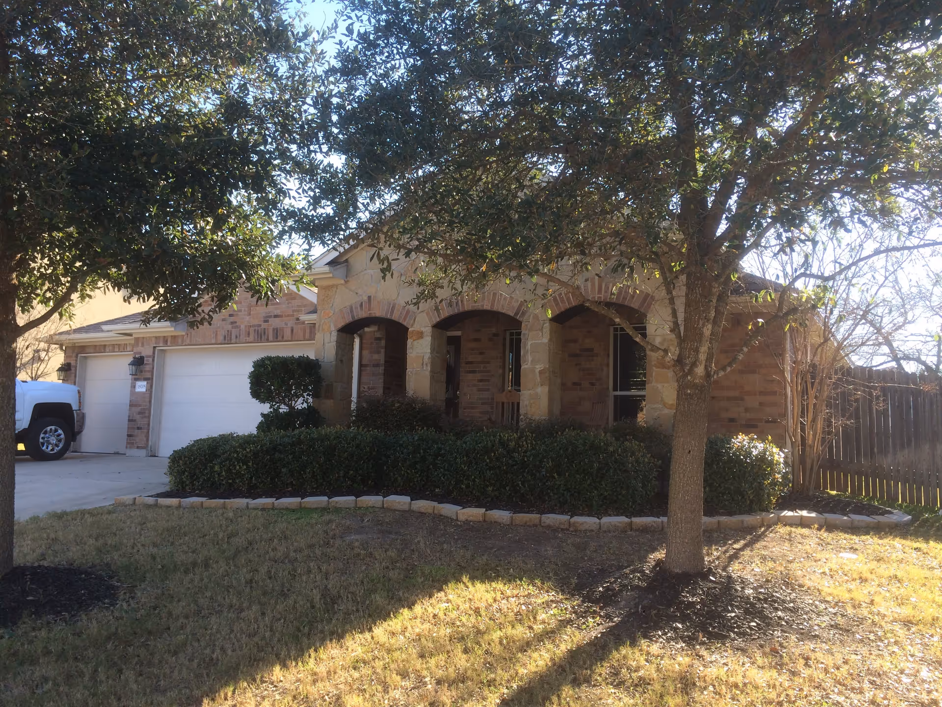 Front exterior view of a single-story brick house with a stone archway porch, two wooden rocking chairs, a double garage, and a driveway with a white truck parked. The yard has grass, bushes, and two trees casting shadows.