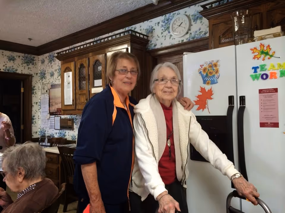 Two elderly women standing in a kitchen area, one using a walker and the other standing beside her with an arm around her shoulder. The kitchen has wooden cabinets, floral wallpaper, a white refrigerator decorated with colorful magnets and notes, and a clock on the wall.