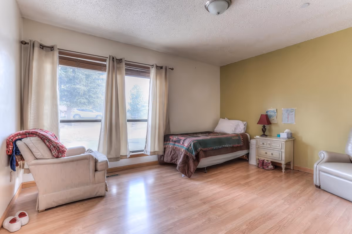 A simple bedroom with a single bed covered with a brown and green blanket, a white pillow, and a small white nightstand with a red lamp and some items on it. There are two children's drawings taped to the yellow wall above the nightstand. The room has light wooden flooring, a beige armchair with a red plaid blanket draped over it, and large windows with beige curtains letting in natural light. A pair of slippers is on the floor near the armchair.