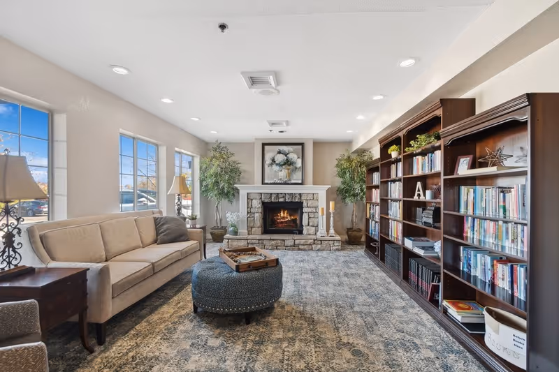 A cozy living room with a beige sofa, a round blue ottoman with a tray on top, two large potted plants, a stone fireplace with a floral painting above it, and a large wooden bookshelf filled with books and decorative items. The room has large windows letting in natural light and a patterned carpet.
