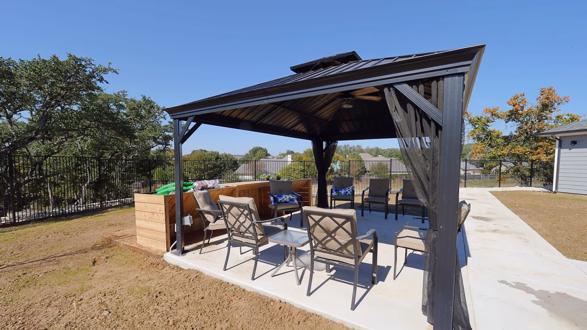 Outdoor seating area under a black metal gazebo with cushioned chairs arranged around a small table, surrounded by a fenced yard with trees and a clear blue sky.