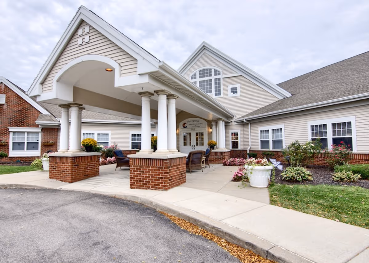 Front exterior view of Peregrine Senior Living at Orchard Park, showing a covered entrance with white columns, brick bases, outdoor seating, and landscaped flower beds under a cloudy sky.
