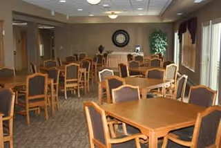Communal dining room with multiple wooden tables and chairs, carpeted floor, a clock on the back wall, and windows along the right side.