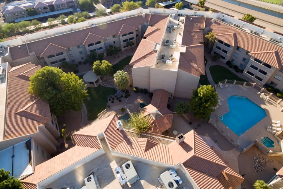 Aerial view of the Grand Court of Mesa senior living facility showing multiple connected buildings with red-tiled roofs surrounding a central courtyard with trees, a gazebo, and a swimming pool area with lounge chairs.