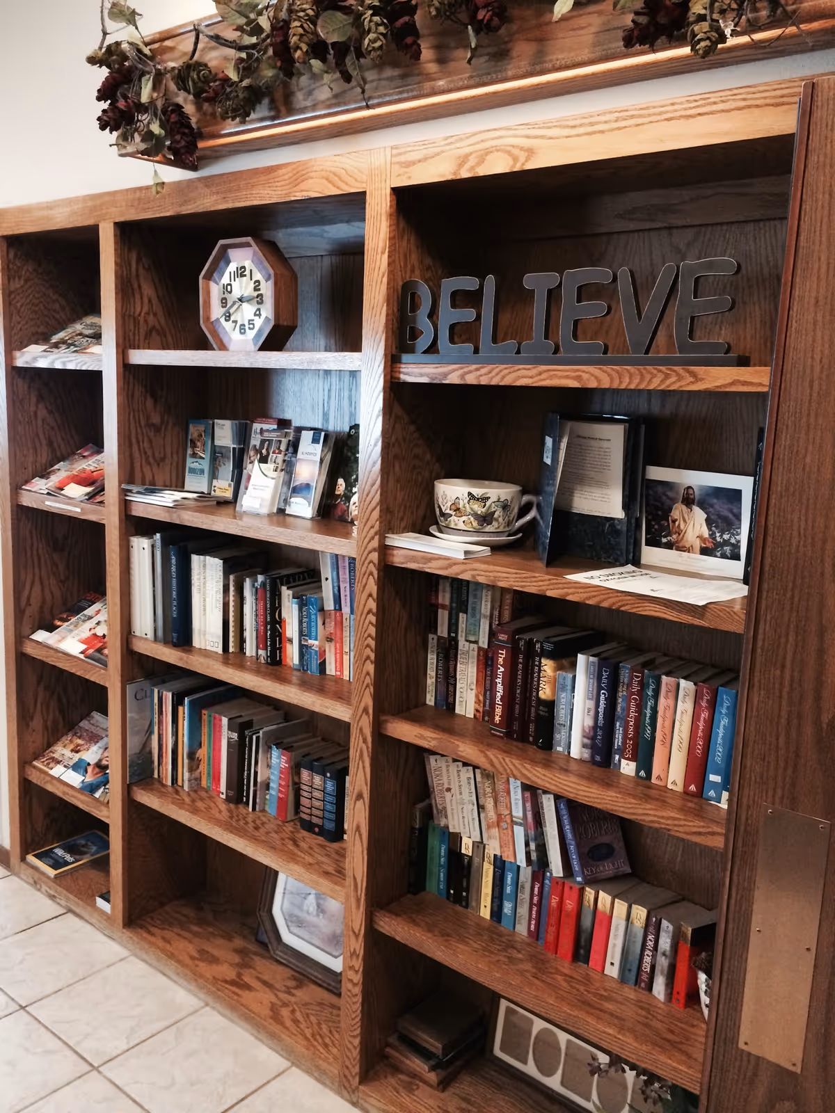 Wooden bookshelf filled with books, pamphlets, a clock, a decorative cup, and a sign that says BELIEVE. There are also some dried flowers hanging above the bookshelf.