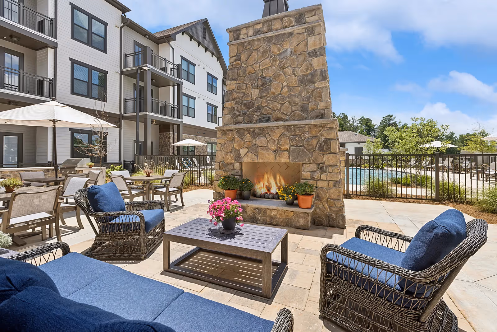 Outdoor patio area at The Phoenix at Opelika featuring a stone fireplace with a fire burning, surrounded by wicker chairs and a sofa with blue cushions, a coffee table with a potted pink flower, several tables with umbrellas, and a fenced swimming pool in the background under a partly cloudy sky.