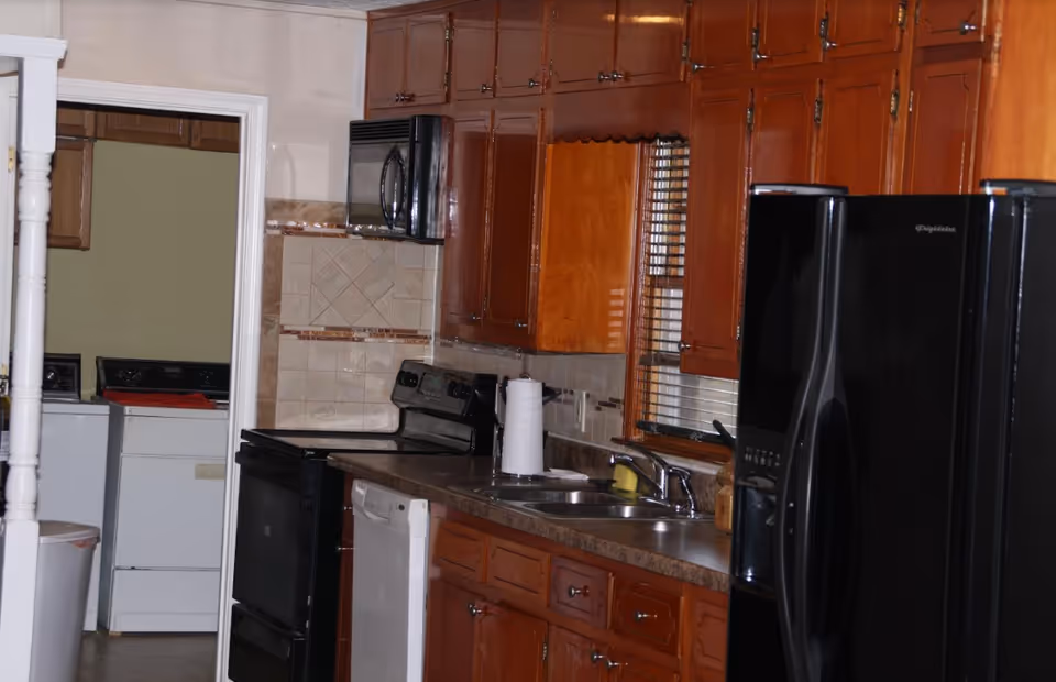 Interior view of a kitchen with wooden cabinets, a black refrigerator, a stove, a microwave, a dishwasher, a double sink with a faucet, and a paper towel holder on the counter. In the background, a laundry area with a washing machine and dryer is visible through an open doorway.