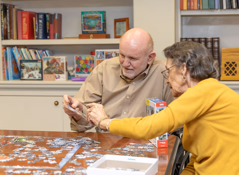 An elderly man and woman sitting at a table working together on assembling a jigsaw puzzle in a room with bookshelves filled with books and puzzles in the background.
