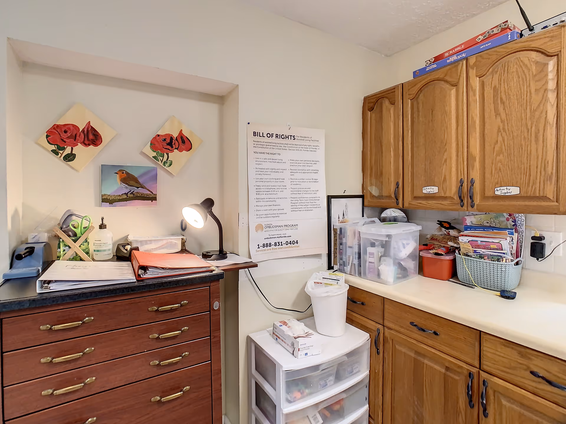 A small workspace area in a senior living facility with wooden cabinets, a countertop holding various containers and supplies, a chest of drawers with a desk lamp, and a wall displaying a Bill of Rights poster and decorative paintings of flowers and a bird.