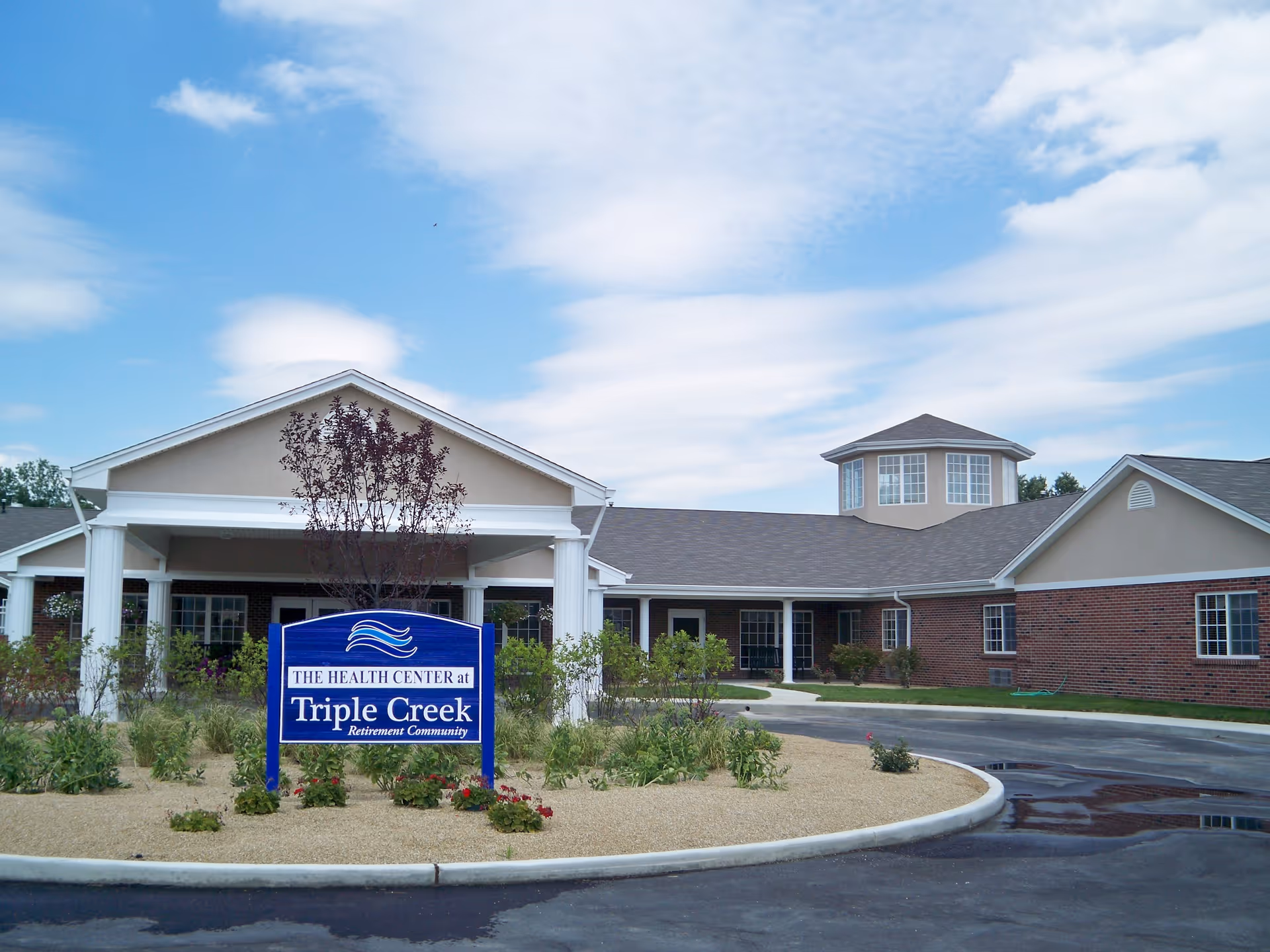 Exterior view of The Health Center at Triple Creek Retirement Community, featuring a single-story brick building with white columns at the entrance, surrounded by landscaping and a circular driveway under a partly cloudy sky.