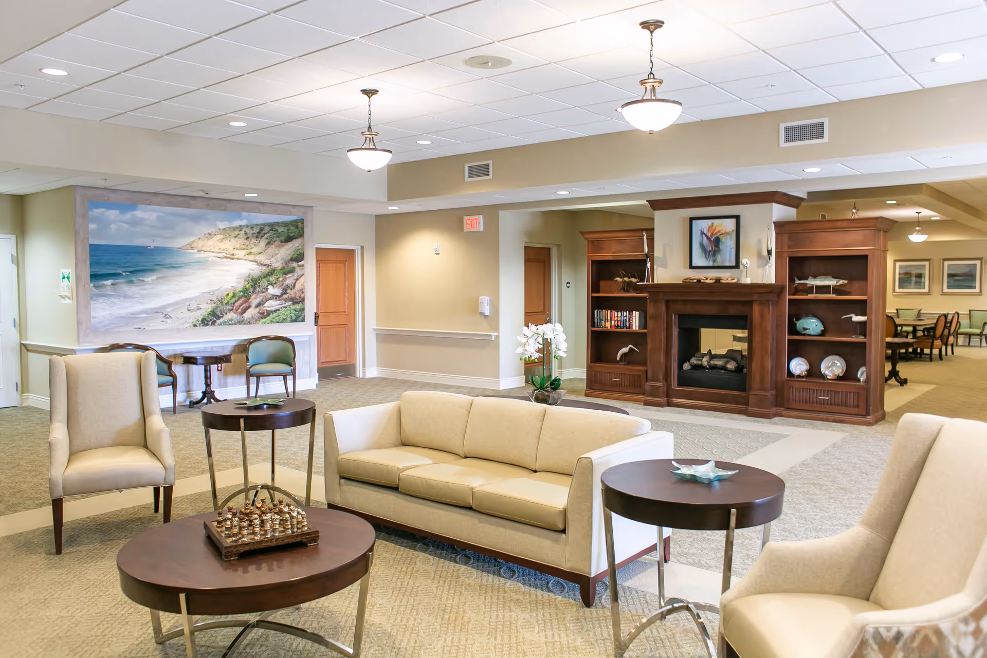 A bright and spacious senior living facility common area with beige sofas and armchairs arranged around dark wooden coffee and side tables. A chess set is on the coffee table. The room features a wooden fireplace with built-in shelves displaying decorative items and books. A large painting of a beach scene is mounted on the wall near two green chairs and a small table. The ceiling has recessed lighting and hanging light fixtures.