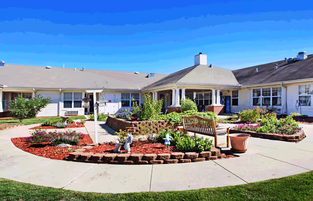Sunny landscaped courtyard with flowerbeds, a wooden bench, and a surrounding single-story senior living building.
