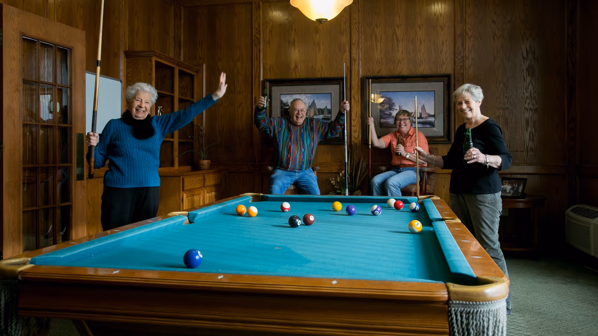 Four elderly people enjoying a game of pool in a wood-paneled room. Three of them are holding pool cues, and two are holding bottles, smiling and celebrating. The room has framed pictures on the walls and a green carpeted floor.