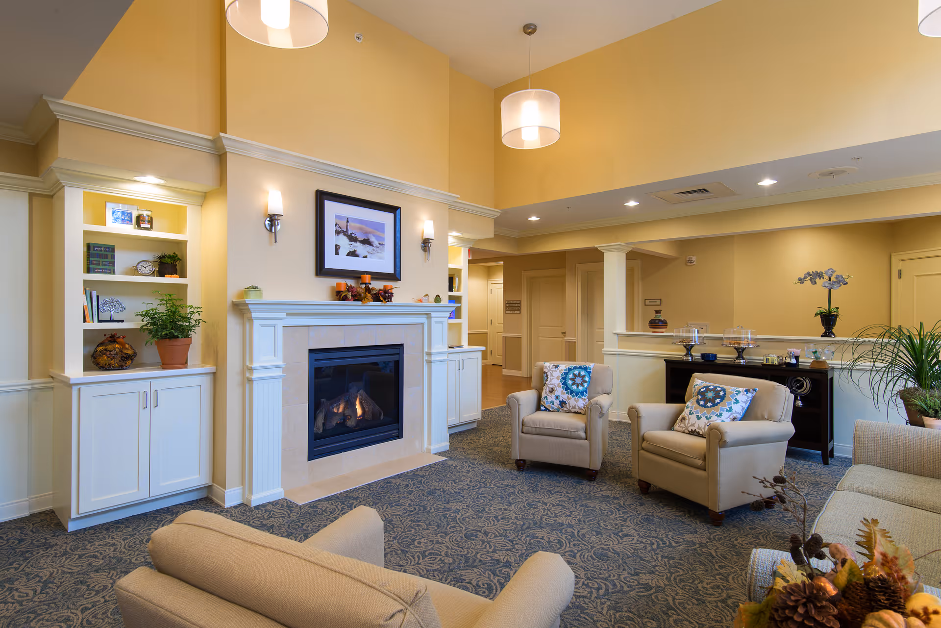 A cozy living room area with beige armchairs and a sofa arranged around a fireplace. The fireplace is framed by white built-in shelves with decorative items and plants. The walls are painted a warm yellow, and the room is lit by ceiling lights and wall sconces. There is a patterned carpet on the floor and a framed picture above the fireplace.