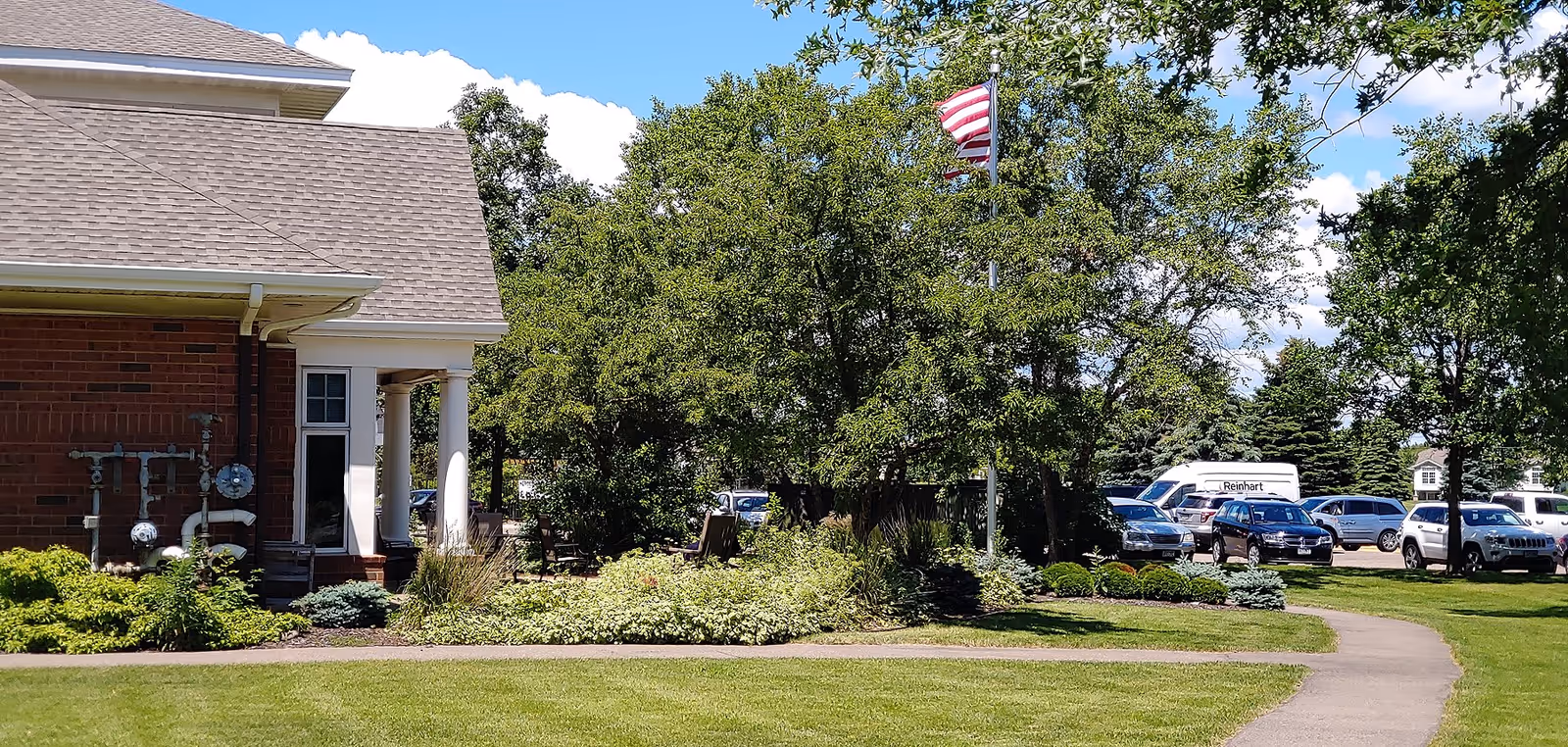 Front exterior of a brick senior living building with white columns, landscaped lawn, an American flag, and parked cars in the background.