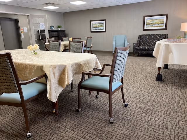 A spacious room with multiple tables covered in beige tablecloths and surrounded by wooden chairs with light blue cushions. The room has carpeted flooring, neutral-colored walls, framed artwork, and a small seating area with a patterned couch and a light blue armchair. There is a lamp on a side table and a cabinet with decorative items in the background.
