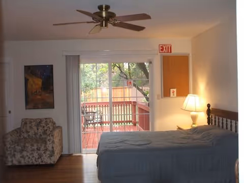 A bedroom with a bed covered in a blue quilt, a floral armchair, a wooden nightstand with a lamp, a ceiling fan, and a sliding glass door leading to an outdoor deck with a table and chairs. There is a painting on the wall and an exit sign above a door.