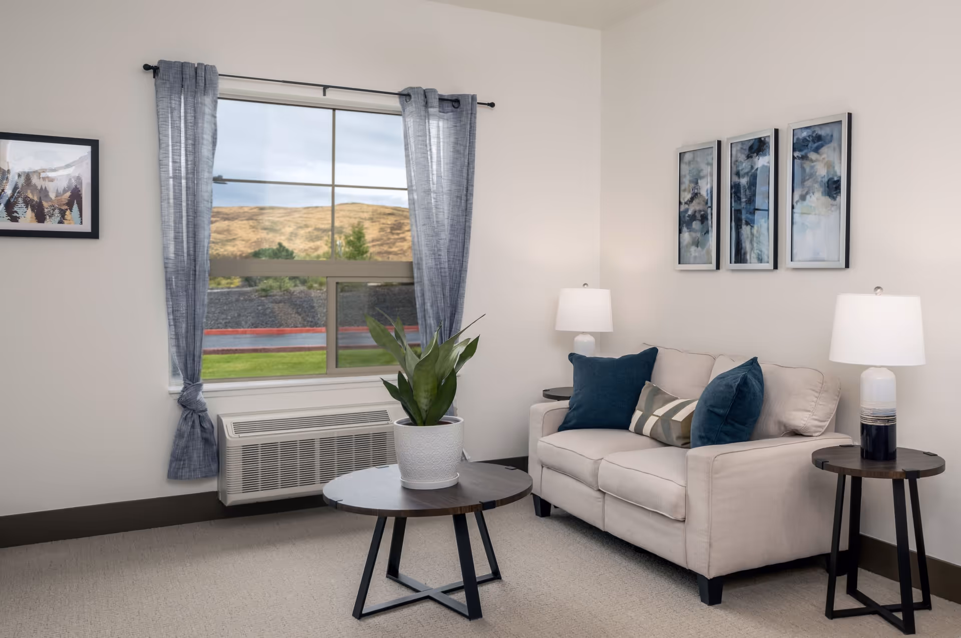 A cozy living room with a beige loveseat adorned with blue and patterned throw pillows. There are two side tables with white lamps on either side of the loveseat. A round wooden coffee table with a potted green plant is in front of the loveseat. The room has a large window with blue curtains tied back, showing a view of a grassy hill outside. The walls are decorated with framed artwork.