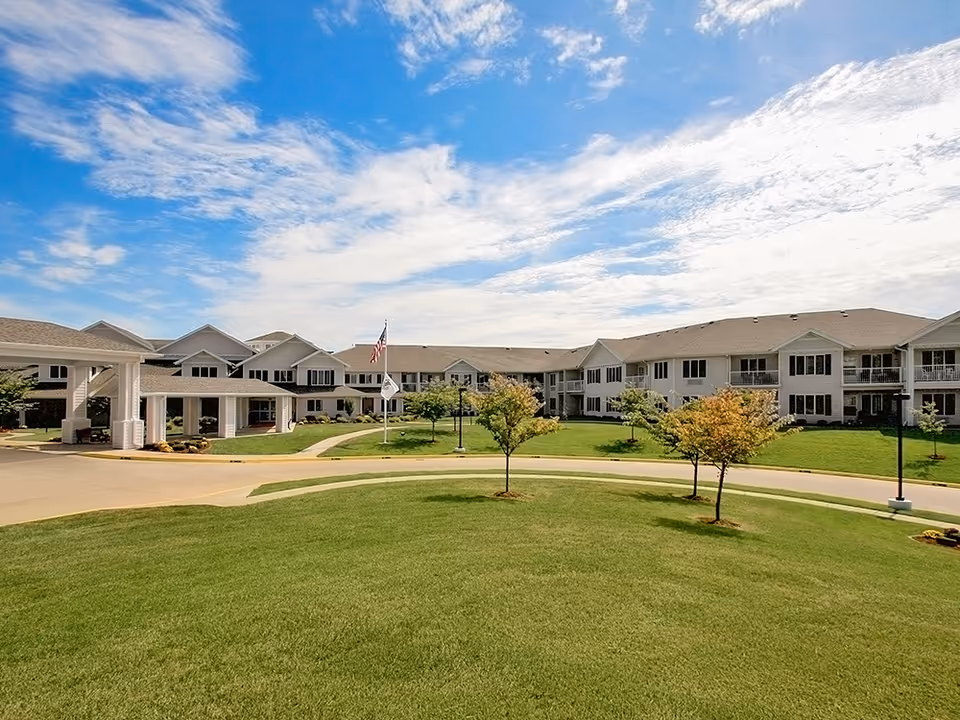 Exterior front view of a two-story senior living building with a large lawn, small trees, flagpoles, and a covered entrance under a blue sky.