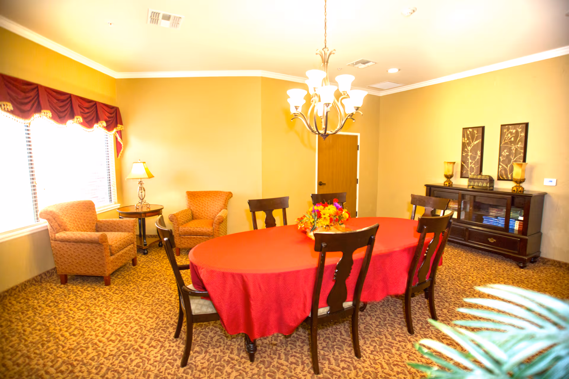 A dining room with a large oval table covered with a red tablecloth and surrounded by six wooden chairs. There is a floral centerpiece on the table. The room has beige walls and carpeted floor with a patterned design. Two upholstered armchairs and a small round table with a lamp are near a window with red valance curtains. A chandelier with multiple lights hangs above the table. Against one wall is a dark wooden cabinet with decorative items and two framed artworks above it.