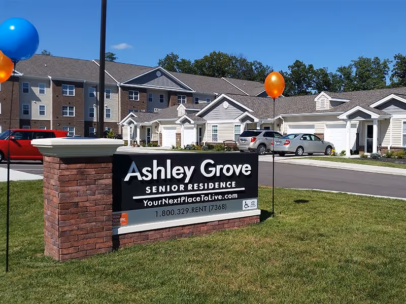 Exterior view of Ashley Grove Senior Residence showing a large sign with the facility name, website, and phone number. Behind the sign are residential buildings, parked cars, and balloons on poles on a sunny day.