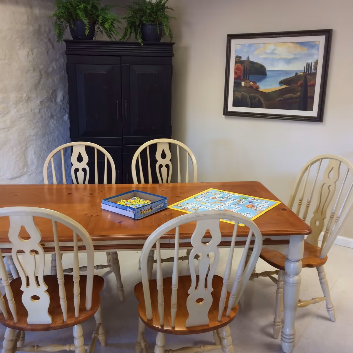 A wooden dining table with six chairs around it. On the table, there is a board game with a blue box and a colorful game board. Behind the table, there is a black cabinet with two potted plants on top. A framed painting of a coastal landscape hangs on the wall.