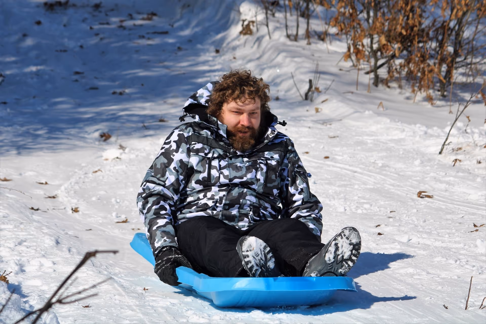 A man wearing a black and white camouflage jacket and black pants is sledding down a snowy hill on a blue sled. The surrounding area is covered in snow with some dry plants and trees visible.