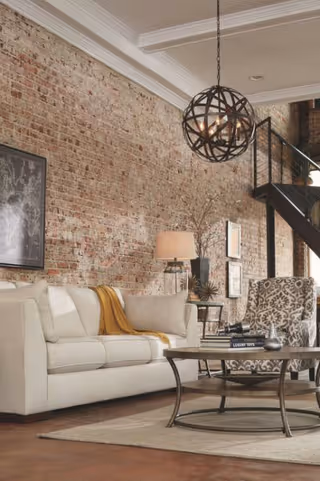 Cozy living room with a white sofa, patterned armchair, round coffee table, exposed brick wall, and a spherical pendant light.