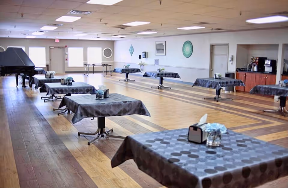 Spacious dining/activity room with multiple small square tables draped in gray polka-dot tablecloths, a grand piano at the rear, and wood-patterned flooring.