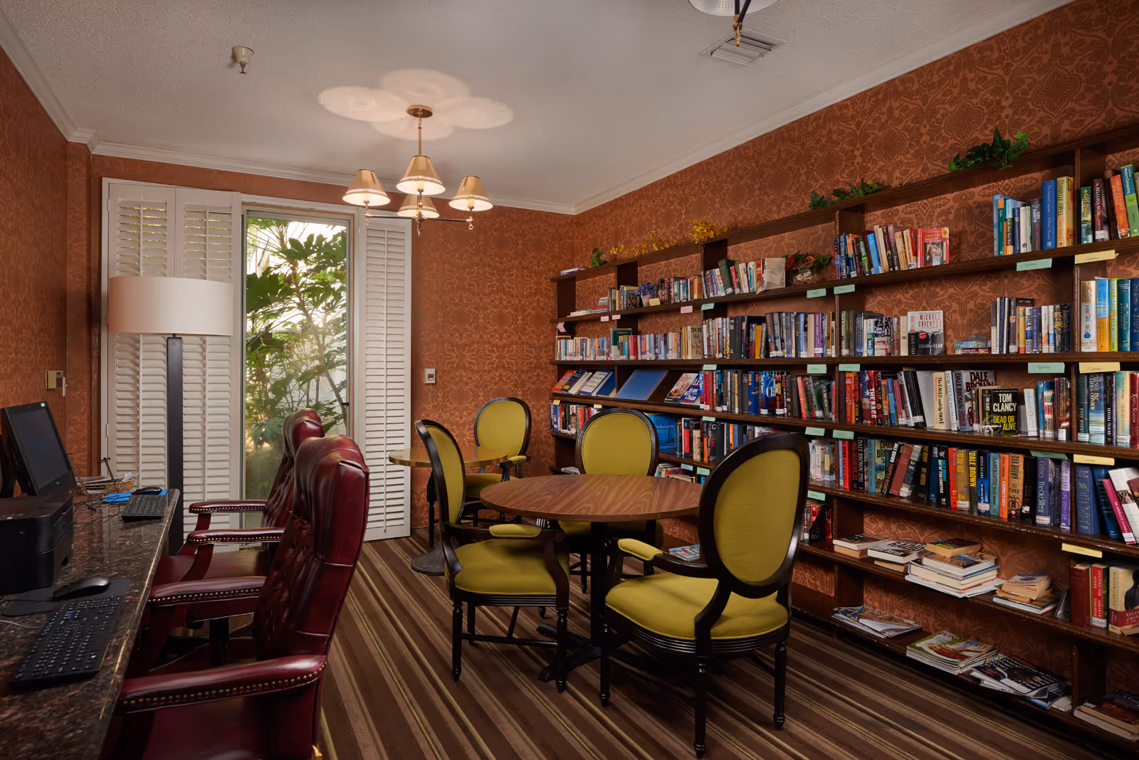 A cozy library room with patterned wallpaper and striped carpet. There are wooden bookshelves filled with books along one wall, a round wooden table surrounded by four green cushioned chairs in the center, and a row of computer stations with red leather chairs along the opposite wall. A window with white plantation shutters lets in natural light, and a ceiling light fixture with three lampshades hangs above.