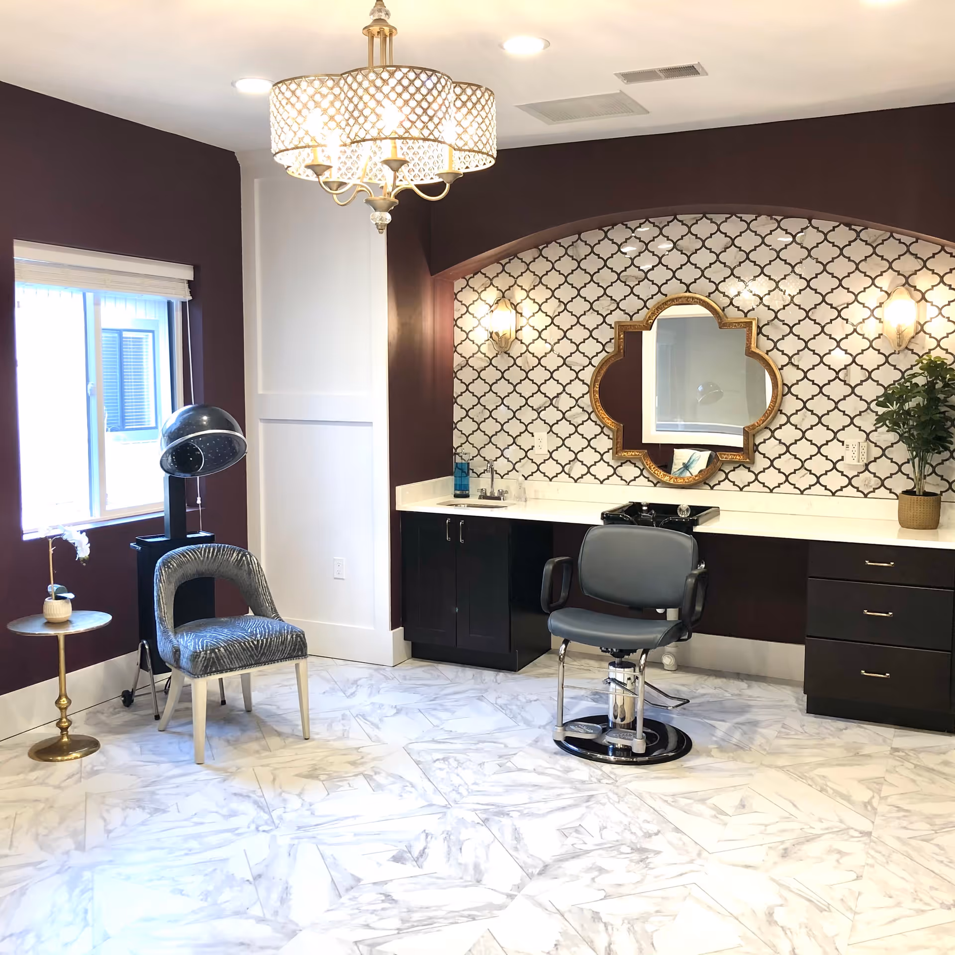 A stylish salon area with a hair washing station, a salon chair, a patterned wall with a decorative mirror, a side chair, a hair dryer, and a small side table with a plant. The room has marble flooring and a chandelier light fixture.