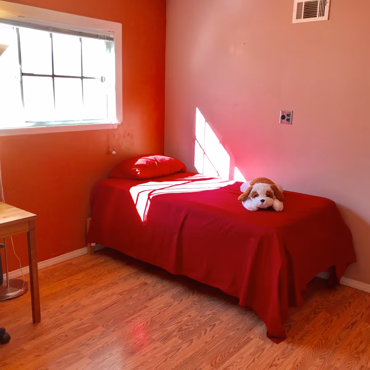 A small bedroom with a single bed covered in a red bedspread and pillow. A stuffed dog toy is placed on the bed. The room has wooden flooring, a window with blinds letting in sunlight, and a small wooden desk with a lamp beside the bed.