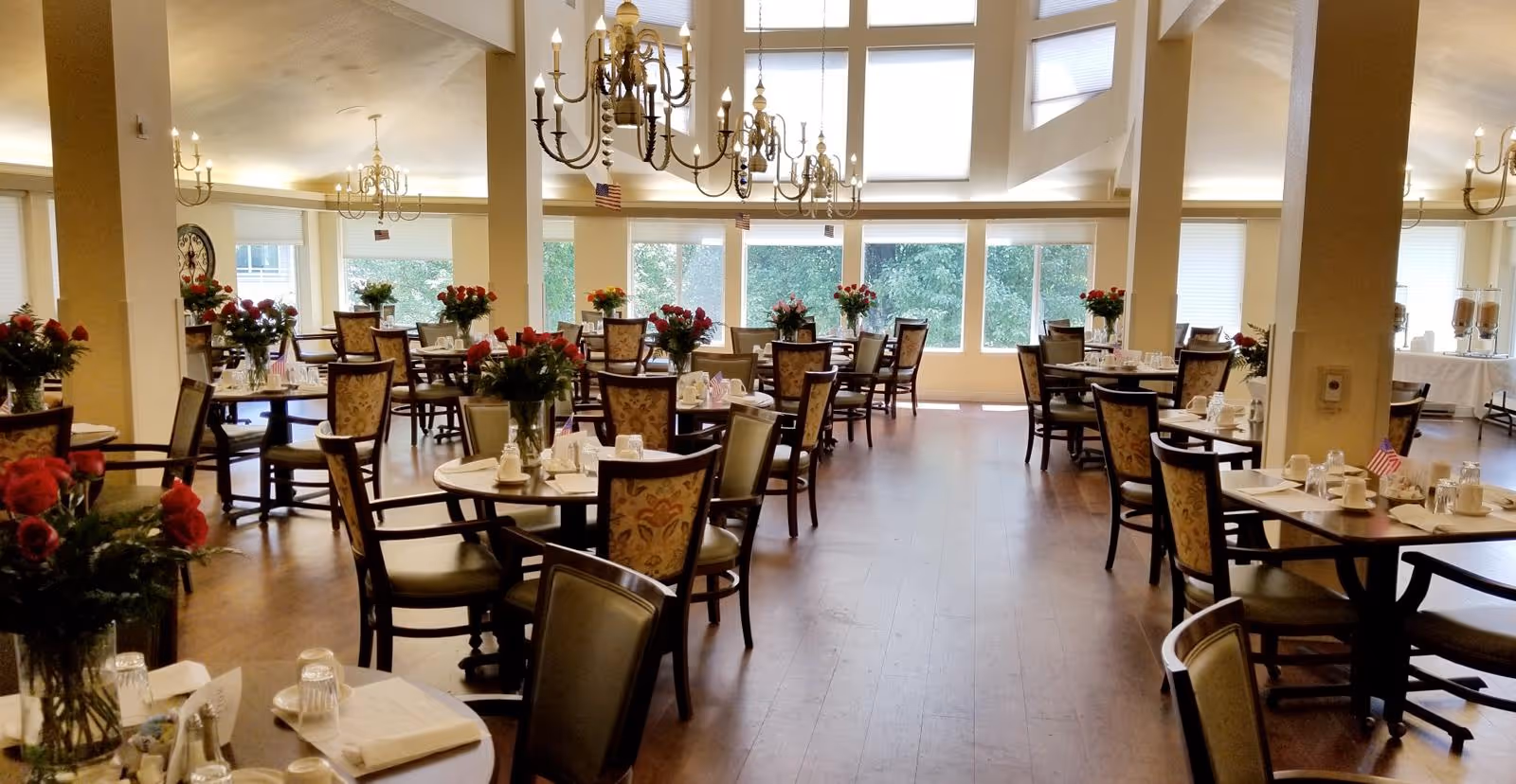 Spacious dining room with multiple round tables and chairs, chandeliers, and floral centerpieces.