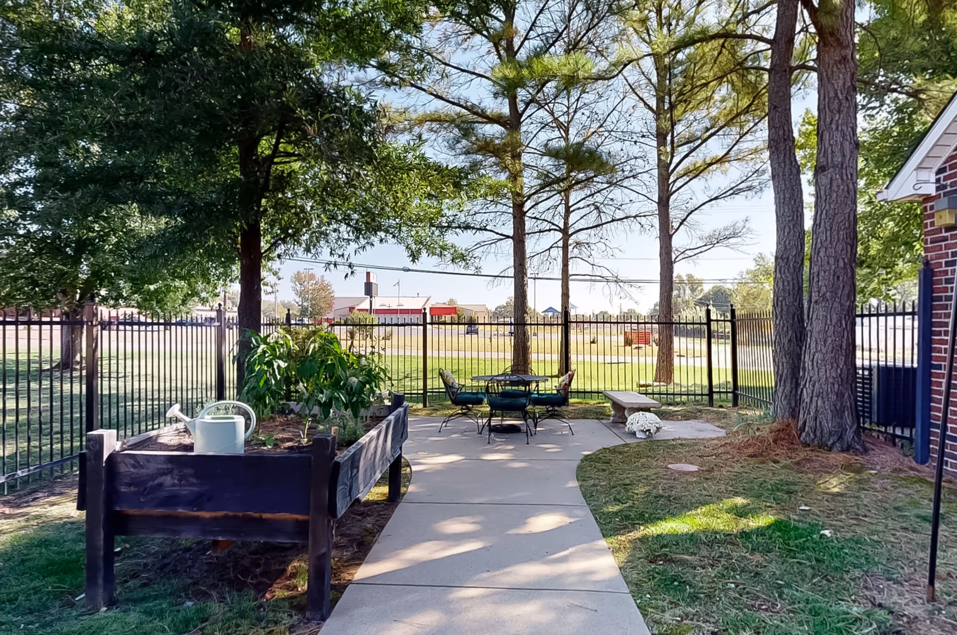 Outdoor patio area at Green Tree Assisted Living featuring a raised garden bed with plants and a watering can, a paved walkway leading to a round table with four chairs, a stone bench, and several trees providing shade. The area is enclosed by a black metal fence with a grassy field and buildings visible in the background.