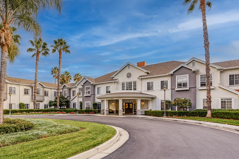 Exterior view of a senior living facility named Gardens At Northridge, featuring a large two-story building with white and gray siding, multiple windows, a covered entrance with columns, palm trees, well-maintained landscaping, and a curved driveway under a partly cloudy sky.