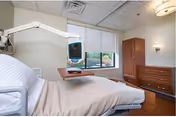 A hospital-style room with a bed featuring white and beige bedding, a mounted medical monitor on an adjustable arm, a window with a view of greenery outside, and wooden furniture including a dresser and a cabinet.