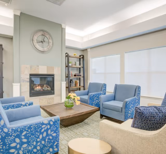 Bright community lounge with blue patterned armchairs arranged around a wooden coffee table in front of a fireplace and wall clock.