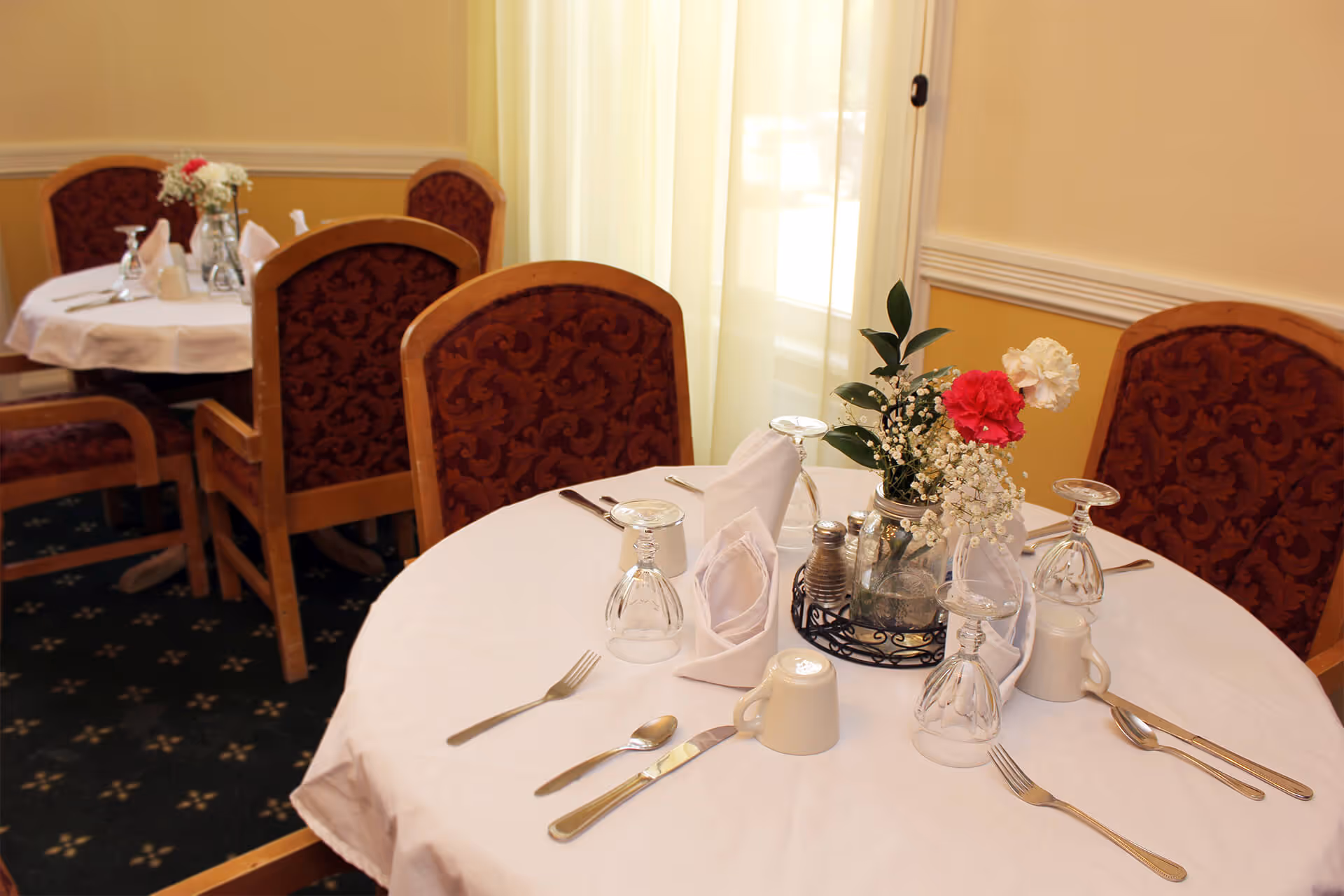 A dining table set for four with white tablecloth, folded white napkins, upside-down wine glasses, silverware, and a centerpiece of flowers in a glass jar. The chairs have wooden frames with red patterned upholstery. Another similarly set table is visible in the background near a window with sheer curtains.